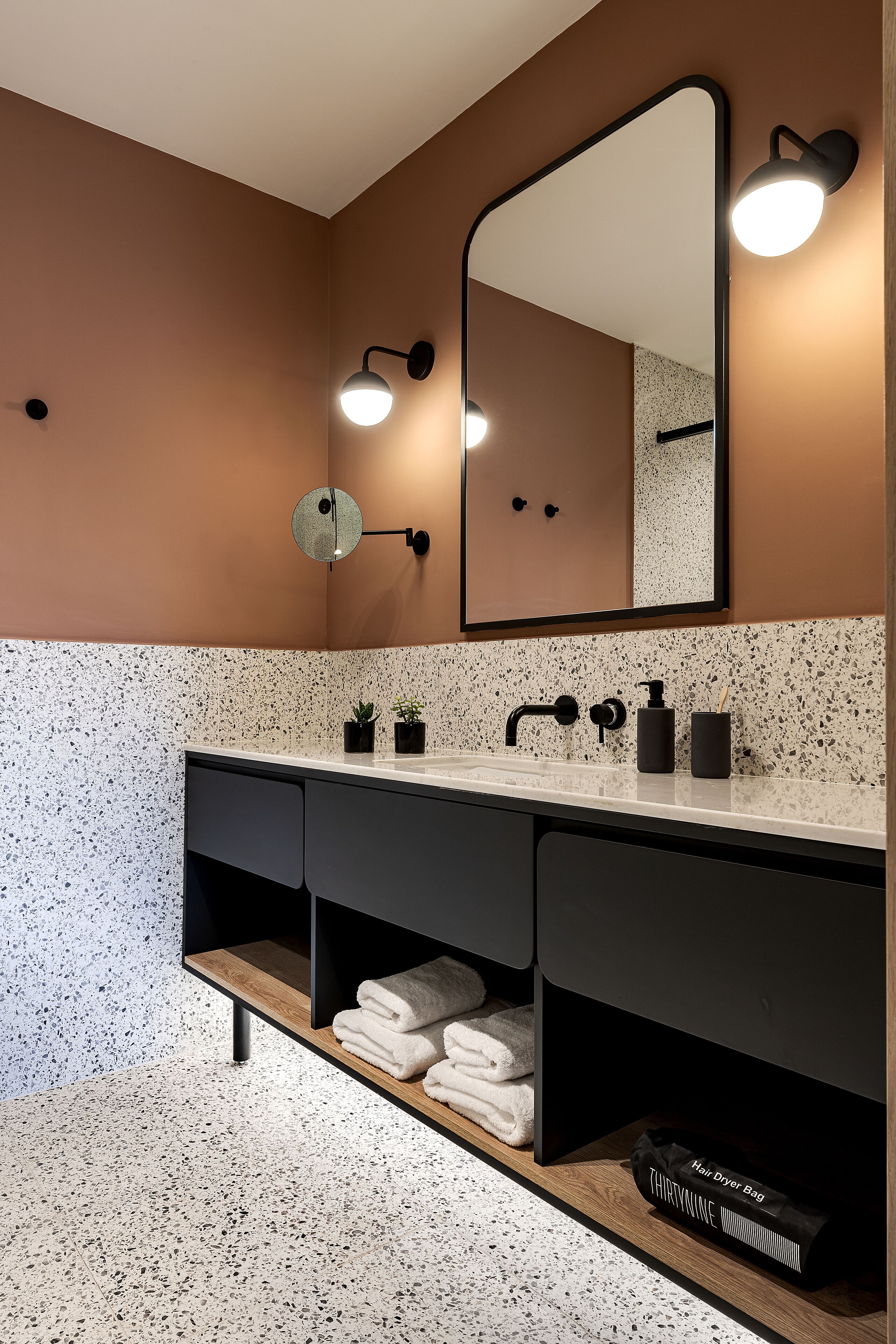 Un baño moderno con piso moteado, un espejo grande, un tocador negro y una bolsa para secador de pelo en el estante.