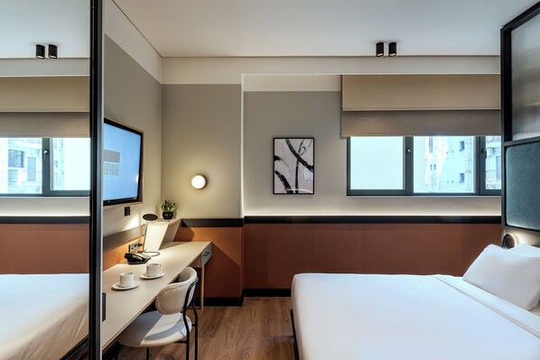 A hotel room with a bed, a desk with a chair, a TV, a wall-mounted lamp, and a window with a view of buildings.