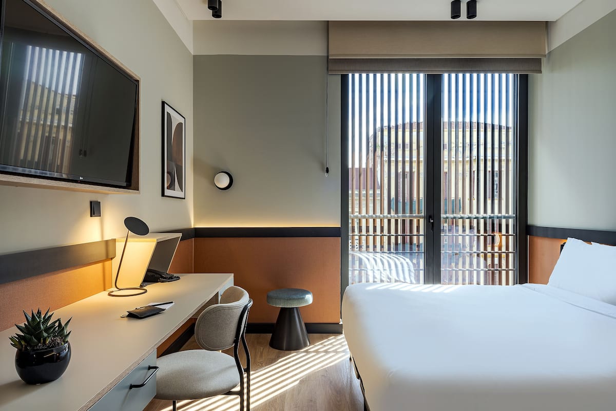 Hotellihuone, jossa on sänky, työpöytä, tuoli, televisio ja ikkunasta avautuva näkymä rakennuksiin.