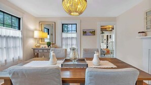 A dining area with a wooden table, grey upholstered chairs, a chandelier, and a view into another room. - Sea Captains Cottage! (Surry)