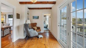 A spacious room with wooden floors, a large window with a view, a chandelier, and a dining area with a table and chairs. - Sea Captains Cottage! (Surry)