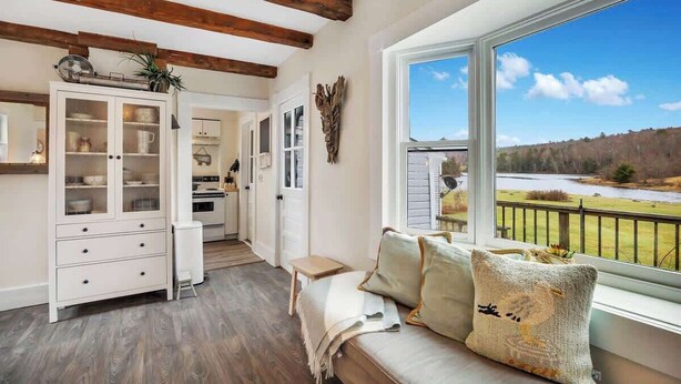 A cozy room with a wooden ceiling, a large window with a view of a lake, a sofa with cushions, and a cabinet with glass doors. - Sea Captains Cottage! (Surry)
