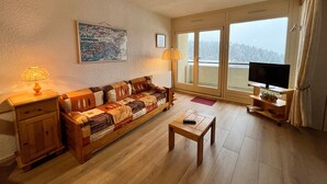 Uma sala de estar acolhedora com sofá de madeira, uma mesinha, televisão em suporte de madeira, abajur e uma janela grande com vista para árvores.