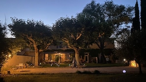 A residential property at dusk with a well-lit front yard, a dining area, and a large tree. - La Bastide des Joncquiers (L'Isle-sur-la-Sorgue)