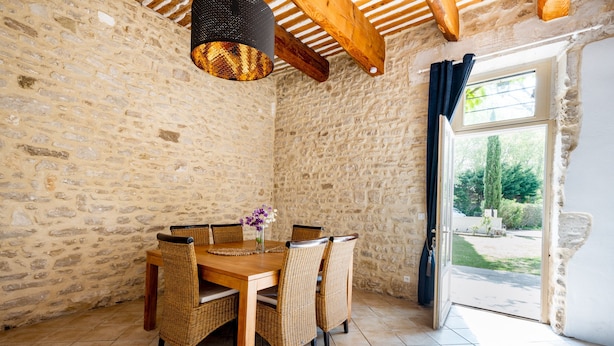 A stone-walled dining area with a wooden table and wicker chairs, a hanging light fixture, and a view through a glass door. - La Bastide des Joncquiers (L'Isle-sur-la-Sorgue)