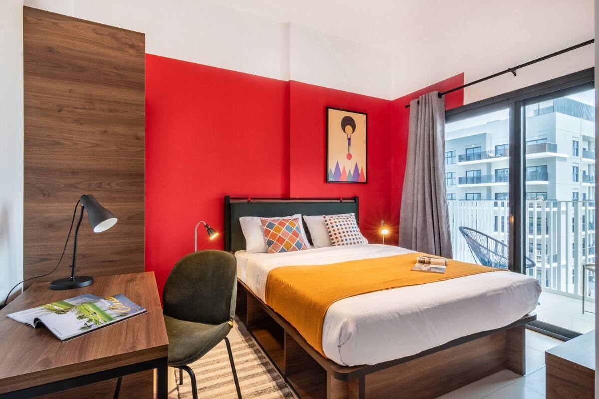 A modern hotel room with a red accent wall, a large bed with multiple pillows, a desk with a lamp, and a view of a cityscape through the window.