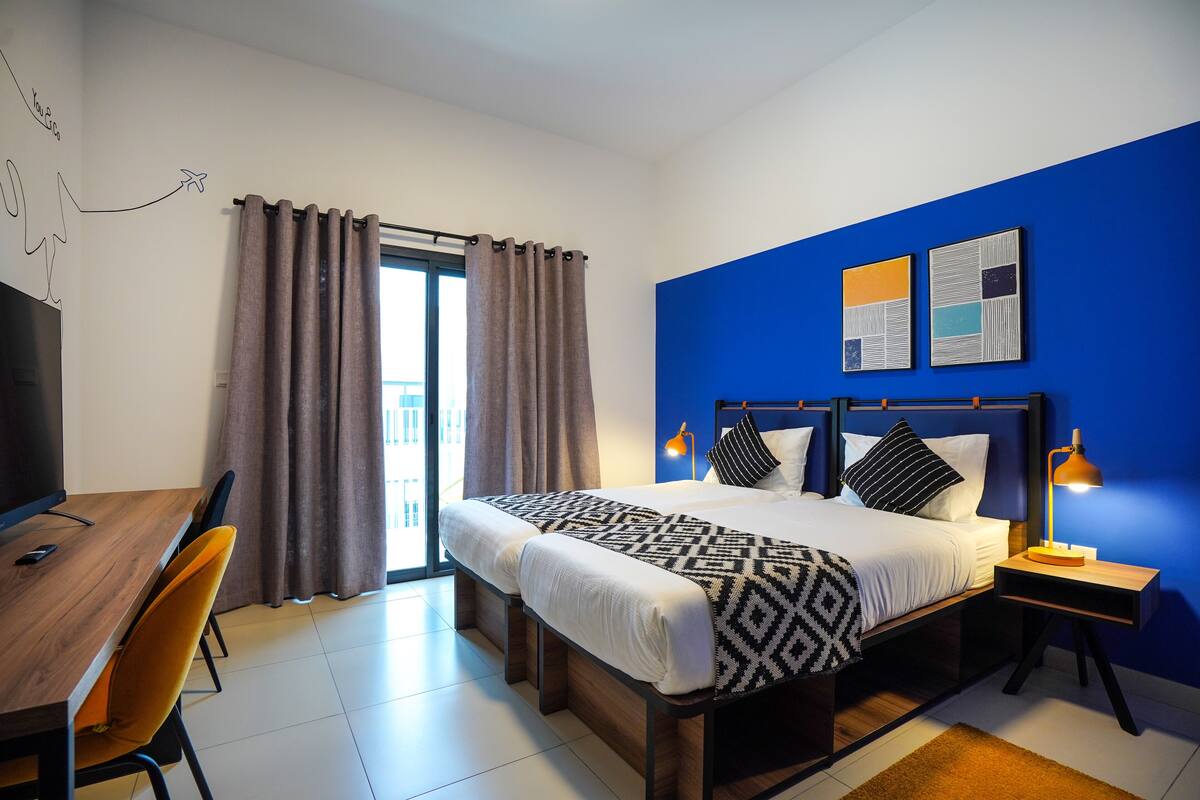 A hotel room with a bed, a desk, a chair, a TV, and a window with curtains.