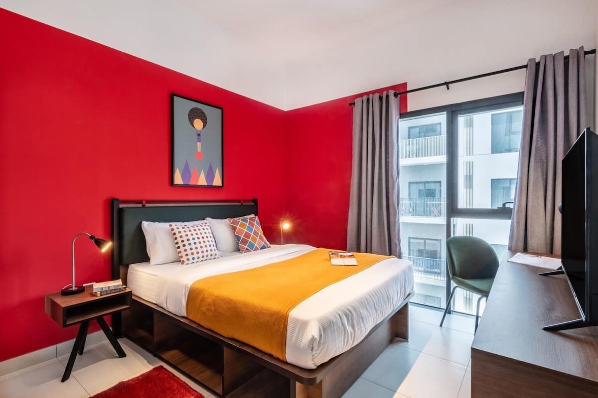 A hotel room with a red wall, a bed with a white and yellow bedspread, a nightstand with a lamp, a desk with a chair, and a large window with curtains.