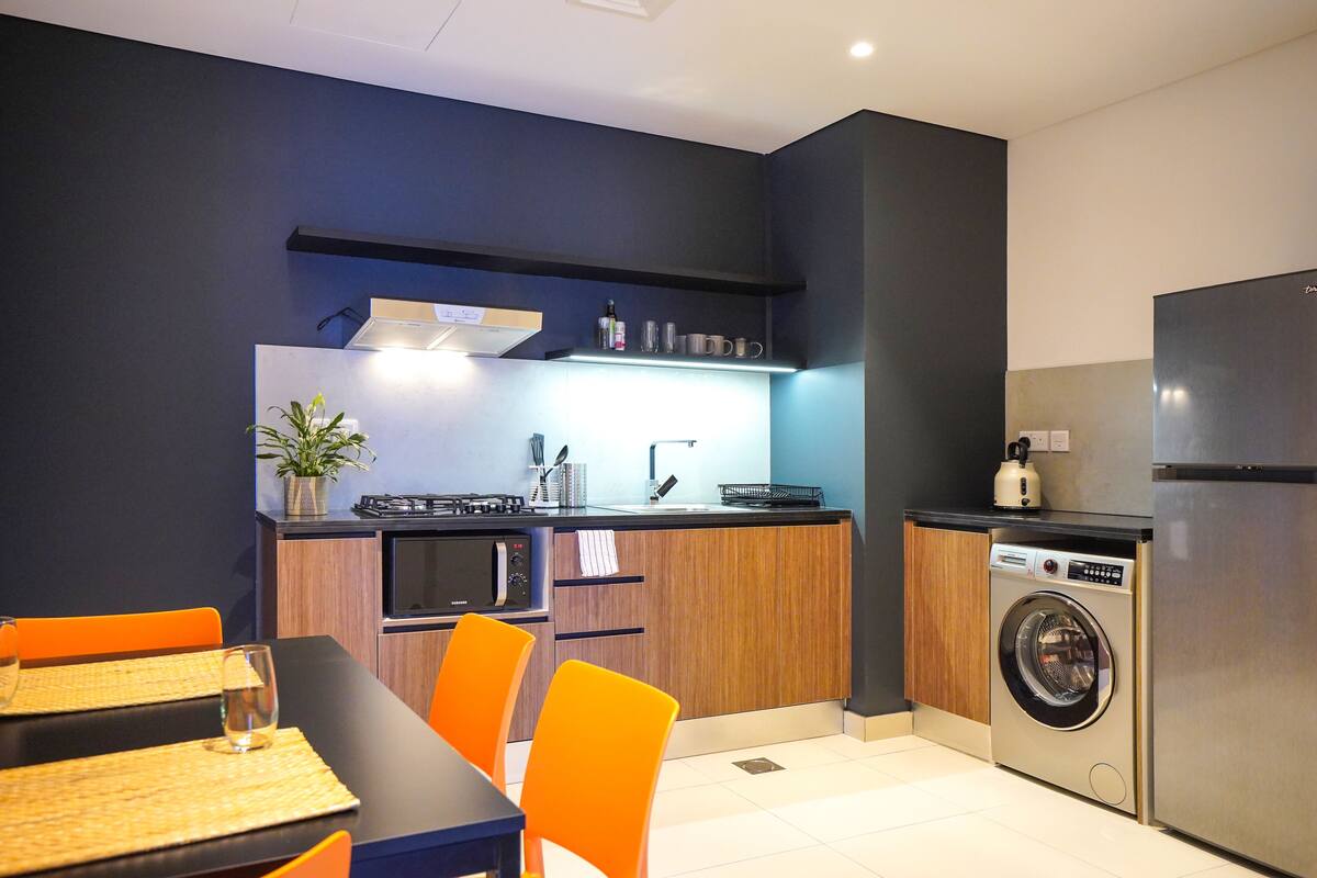 A compact kitchen with a dining area, including a table with chairs, a washing machine, and a refrigerator.