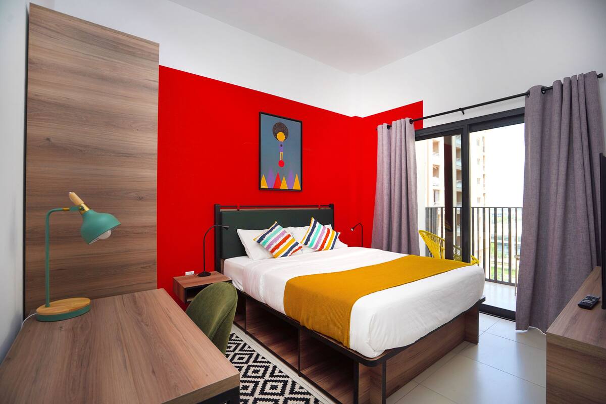 A modern hotel room with a red accent wall, a bed with colorful pillows, a yellow throw, a green chair, a desk with a lamp, and a view of the city through large windows.