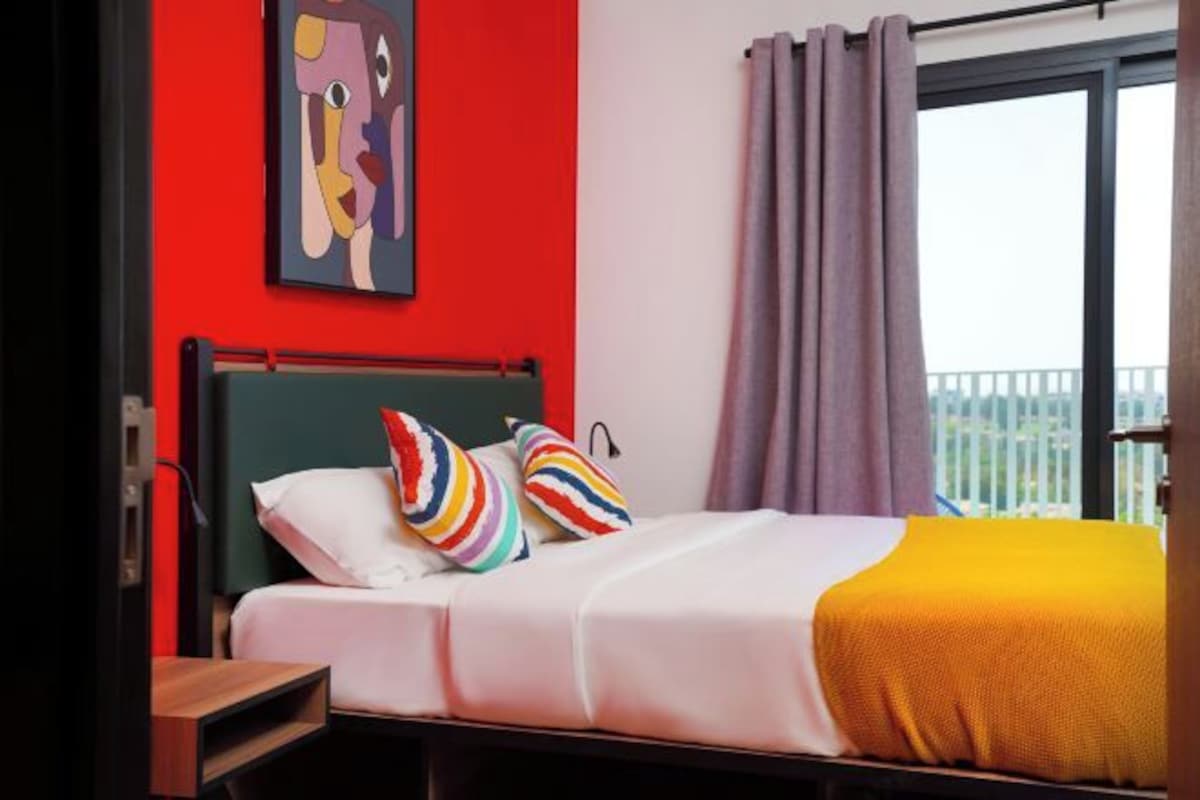 A hotel room with a bed, colorful pillows, a red wall, and a window with curtains.