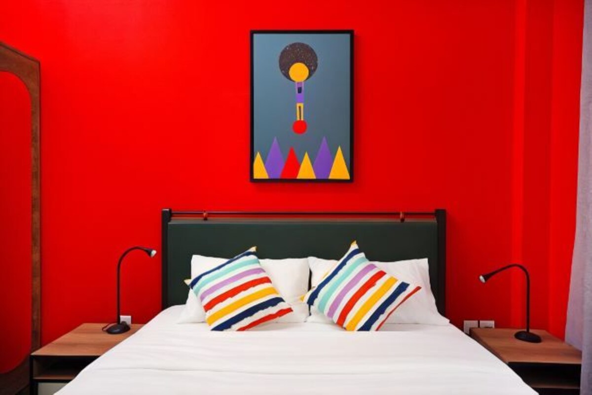 A bed with white bedding and colorful striped pillows, a dark green headboard, and a framed abstract artwork above it.