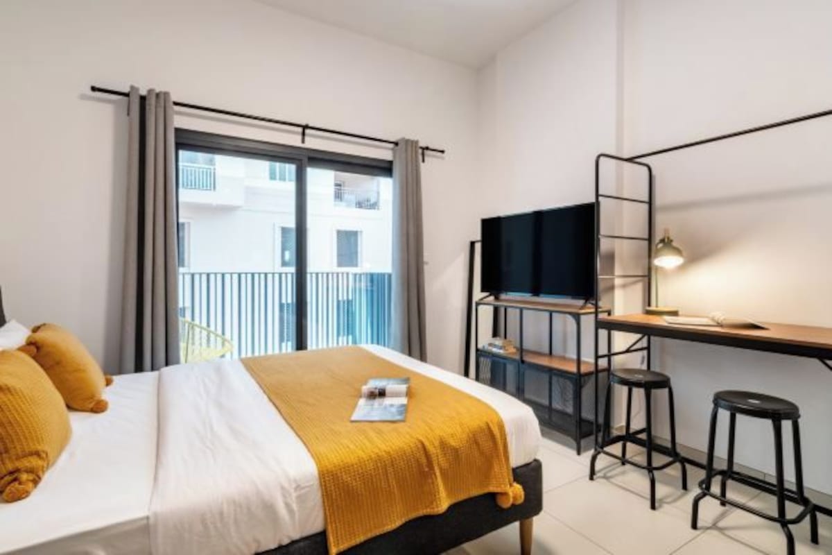 A modern hotel room with a bed, a desk with a chair, a TV, and a balcony view.