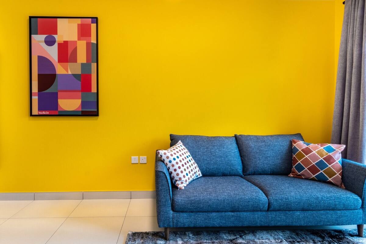 A room with a blue sofa, two patterned pillows, a framed abstract artwork, and a yellow wall.