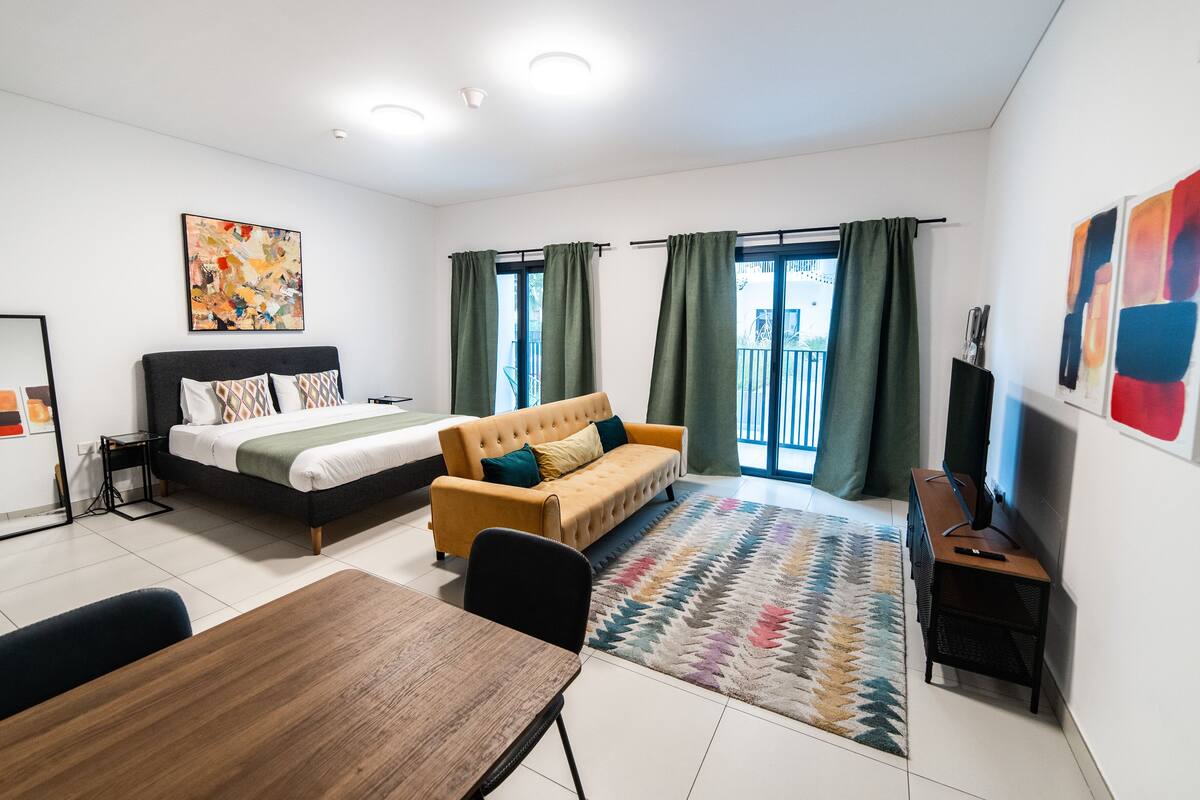 A modern hotel room with a bed, sofa, dining table, and a view of the outdoors.