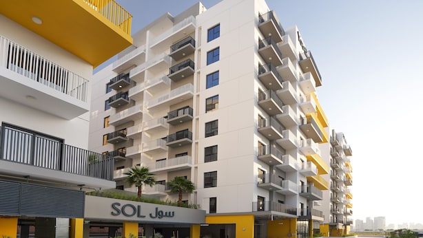 En modern flervåningsbyggnad med bostäder, balkonger, en skylt med texten ”SOL Jou” och ett välanlagt område med växter.
