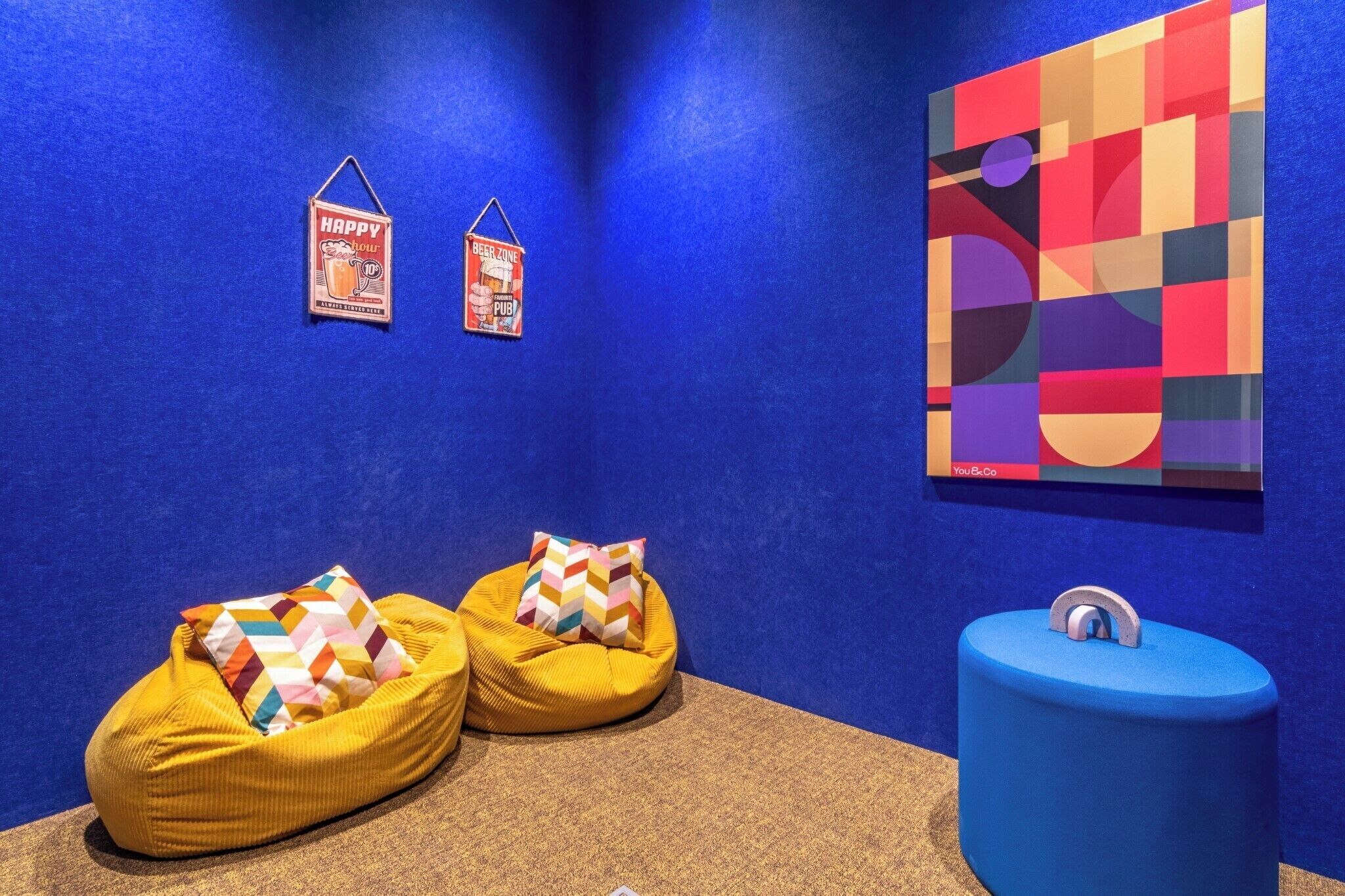A room with blue walls, two bean bags with colorful pillows, a blue trash bin, and two wall decorations.
