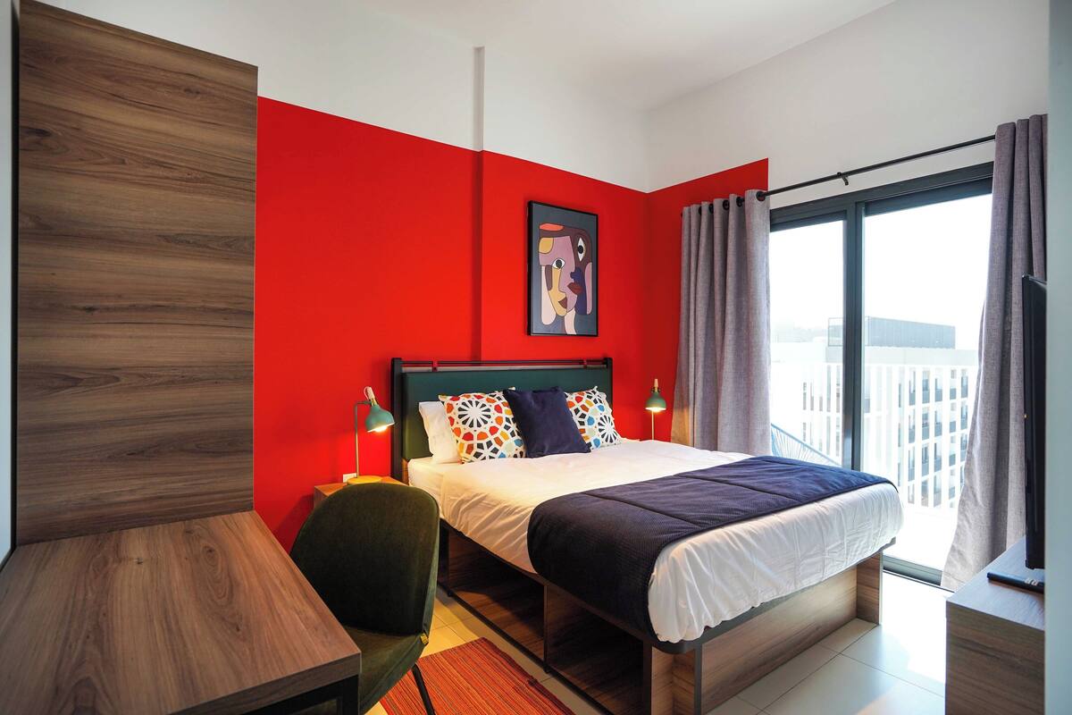 A modern hotel room with a wooden desk, a bed with patterned pillows, and a large window with curtains.