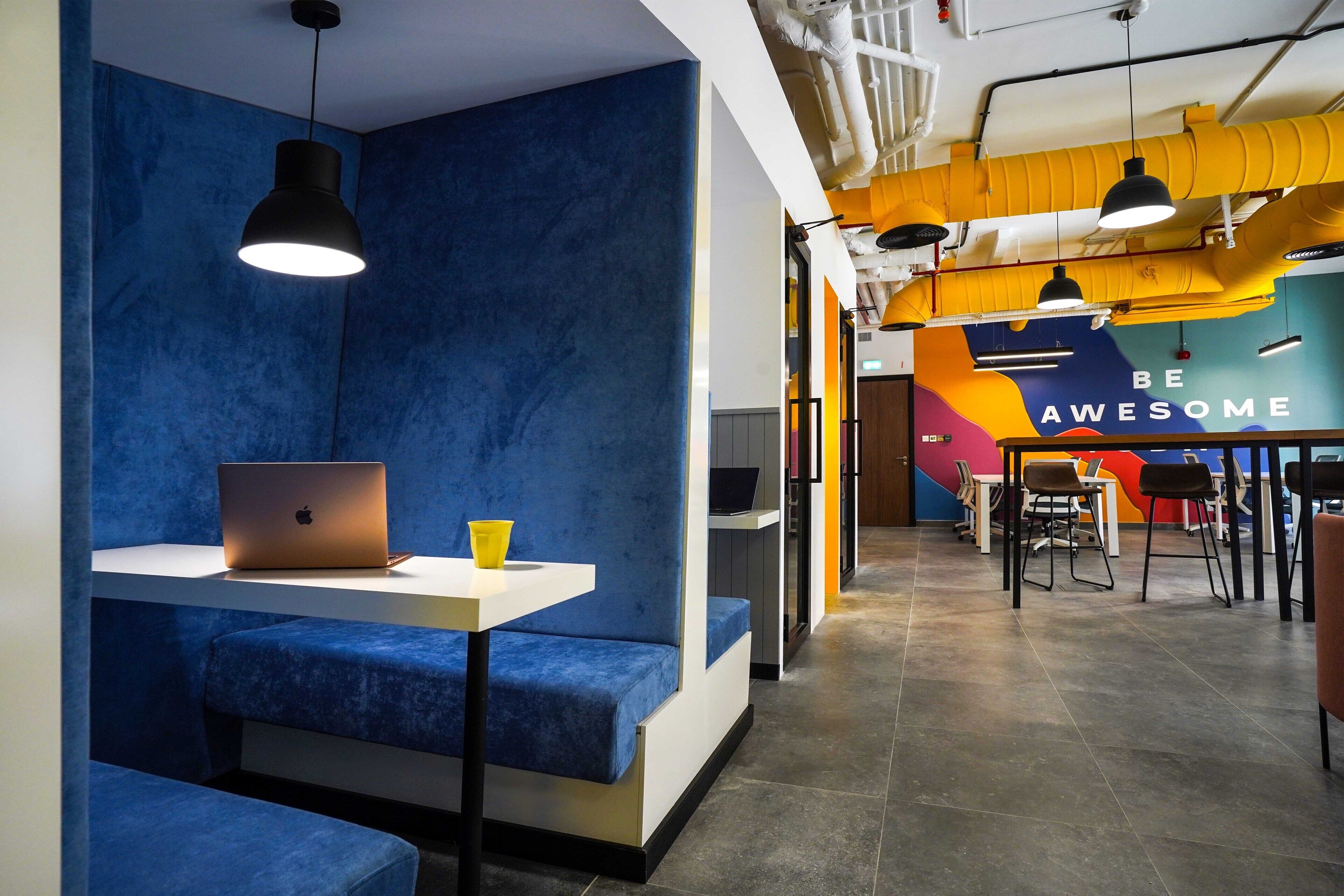 A modern office space with blue seating areas, a white table with a laptop, and a motivational wall with the phrase "BE AWESOME".