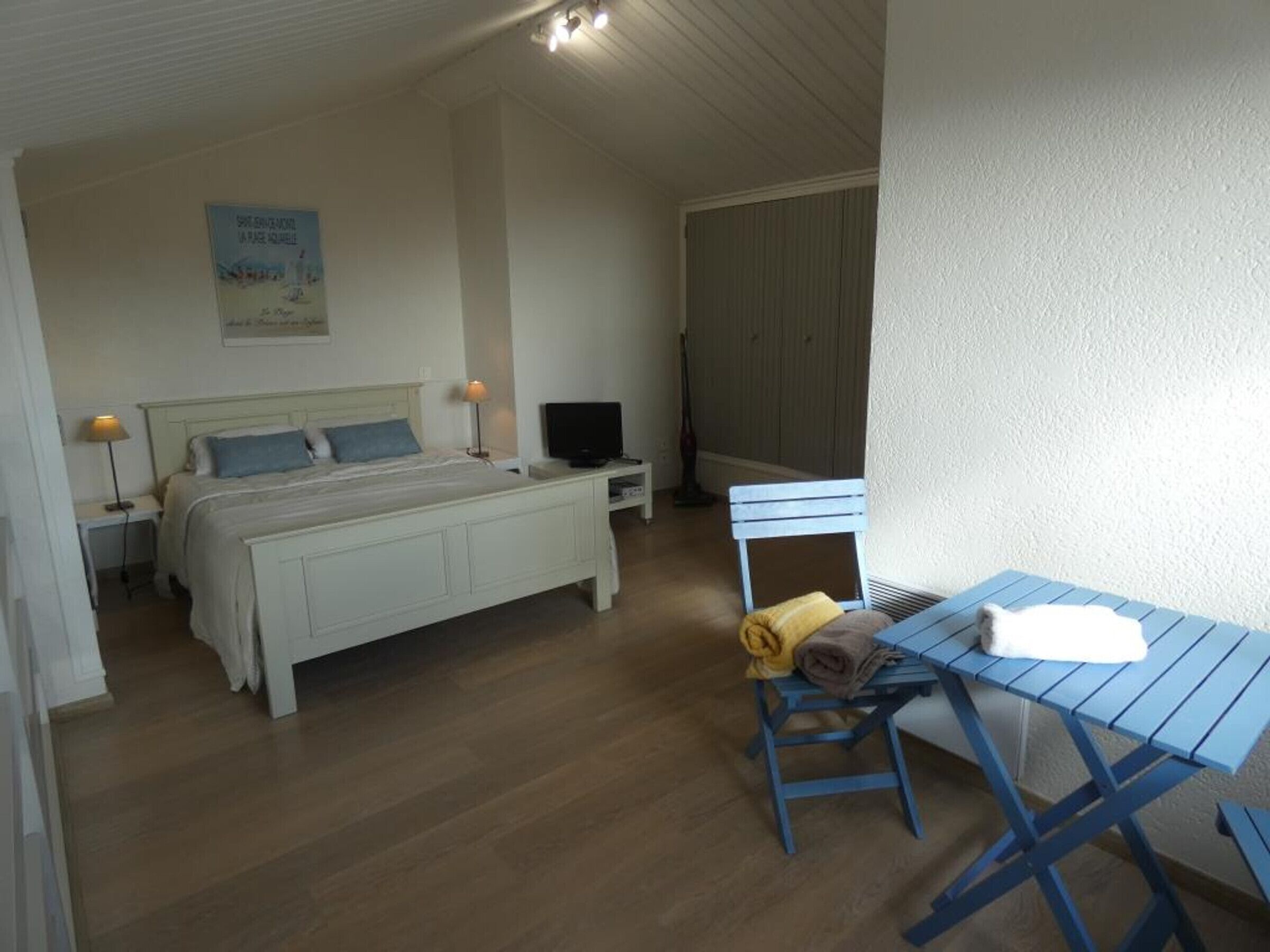 A bedroom with a bed, bedside tables, a TV, a chair with towels, and a poster on the wall.