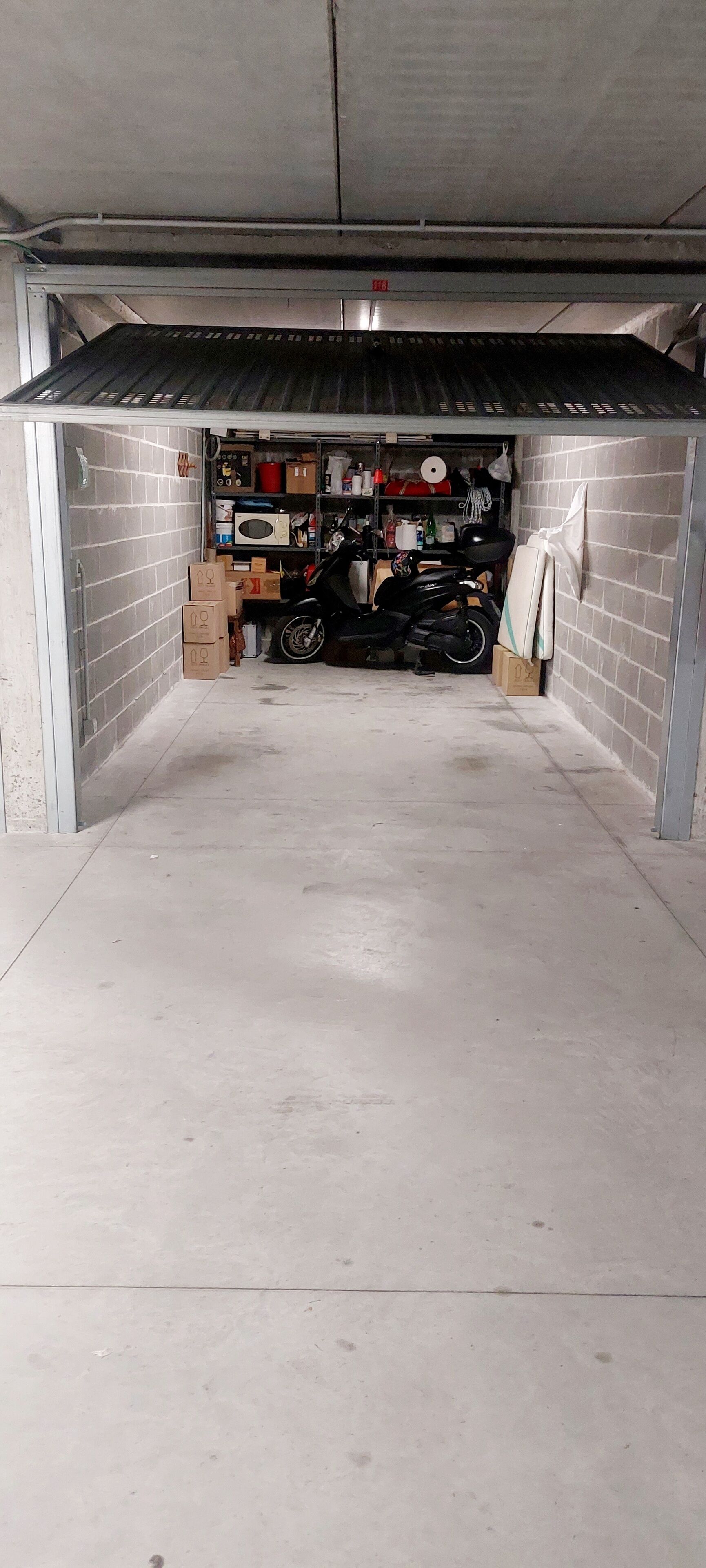 Un garage avec un sol en béton, un toit en métal et un mur en briques. À l’intérieur, on trouve des étagères remplies de divers objets, une moto et quelques cartons.