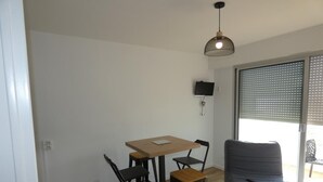 A modern living room with a wooden dining table, two black chairs, a blue sofa, and a large window with blinds. - Seafront Studio at Notre-Dame-de-Monts 2 Pers (Notre-Dame-de-Monts)