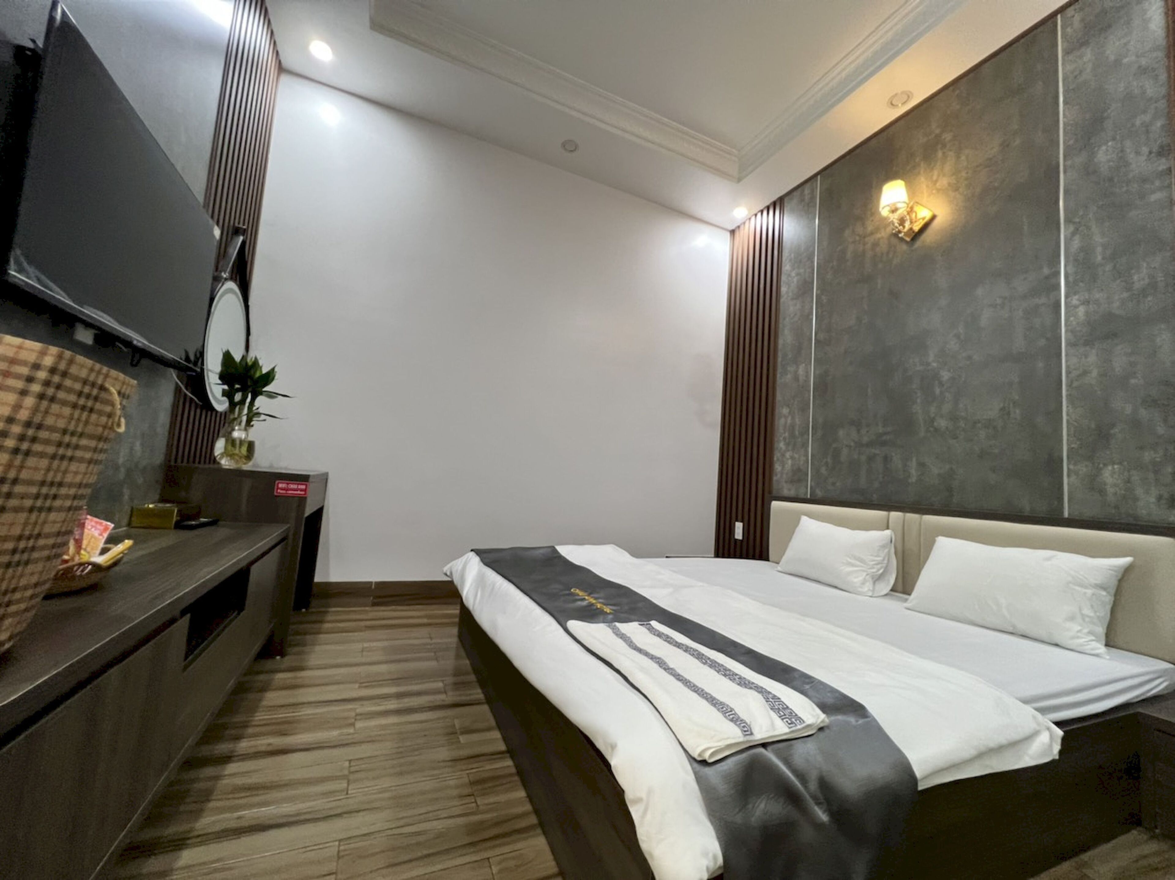 A modern hotel room with a large bed, a flat-screen TV, a wooden dresser, and a wall-mounted light fixture.