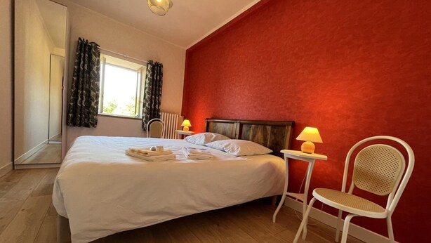 A hotel room with a large bed, a red accent wall, a window with curtains, a bedside table with a lamp, and a chair. - Golf getaway and relaxation at Domaine de Bisseret (Lavault-Sainte-Anne)