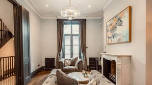 Un salon spacieux avec un grand canapé, une table basse et une cheminée. Un escalier se trouve sur la gauche, ainsi qu’une grande fenêtre avec des rideaux.