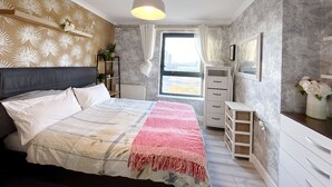 A bedroom with a bed, a bedside table, a dresser, a radiator, a window with curtains, and a hanging light fixture. - Canary Wharf two Bedroom Apartment (London)