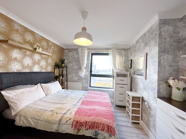 Une chambre à coucher comprenant un lit, une table de chevet, une commode, un radiateur, une fenêtre avec des rideaux et un luminaire suspendu.