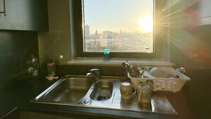 A kitchen with a stainless steel sink, a window with a view of a cityscape, and a dish rack with utensils. - Canary Wharf two Bedroom Apartment (London)
