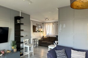 Un salon moderne avec un canapé gris, un fauteuil blanc et une petite table agrémentée d’objets décoratifs. Un téléviseur est fixé au mur, un escalier mène à l’étage supérieur et une fenêtre est dotée de rideaux.
