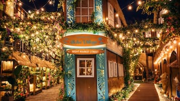A quaint building with a sign reading 'Aloha Village' and 'Night Market Street,' surrounded by lush greenery and string lights.