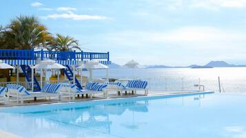 A poolside area with lounge chairs and umbrellas, a blue and white structure, and a view of the sea.