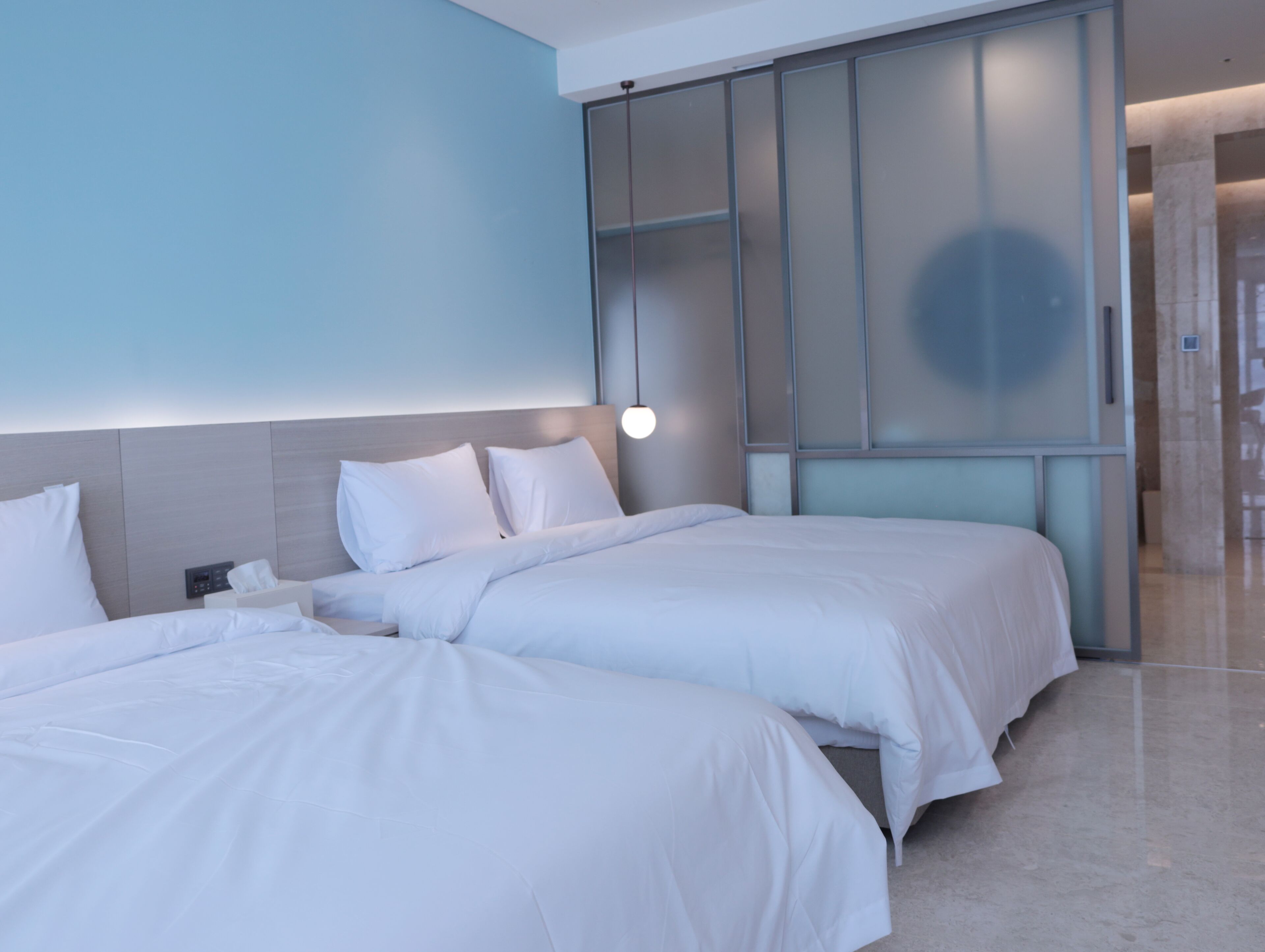 A modern hotel room with two beds, a large mirror, and a light fixture above the bed.
