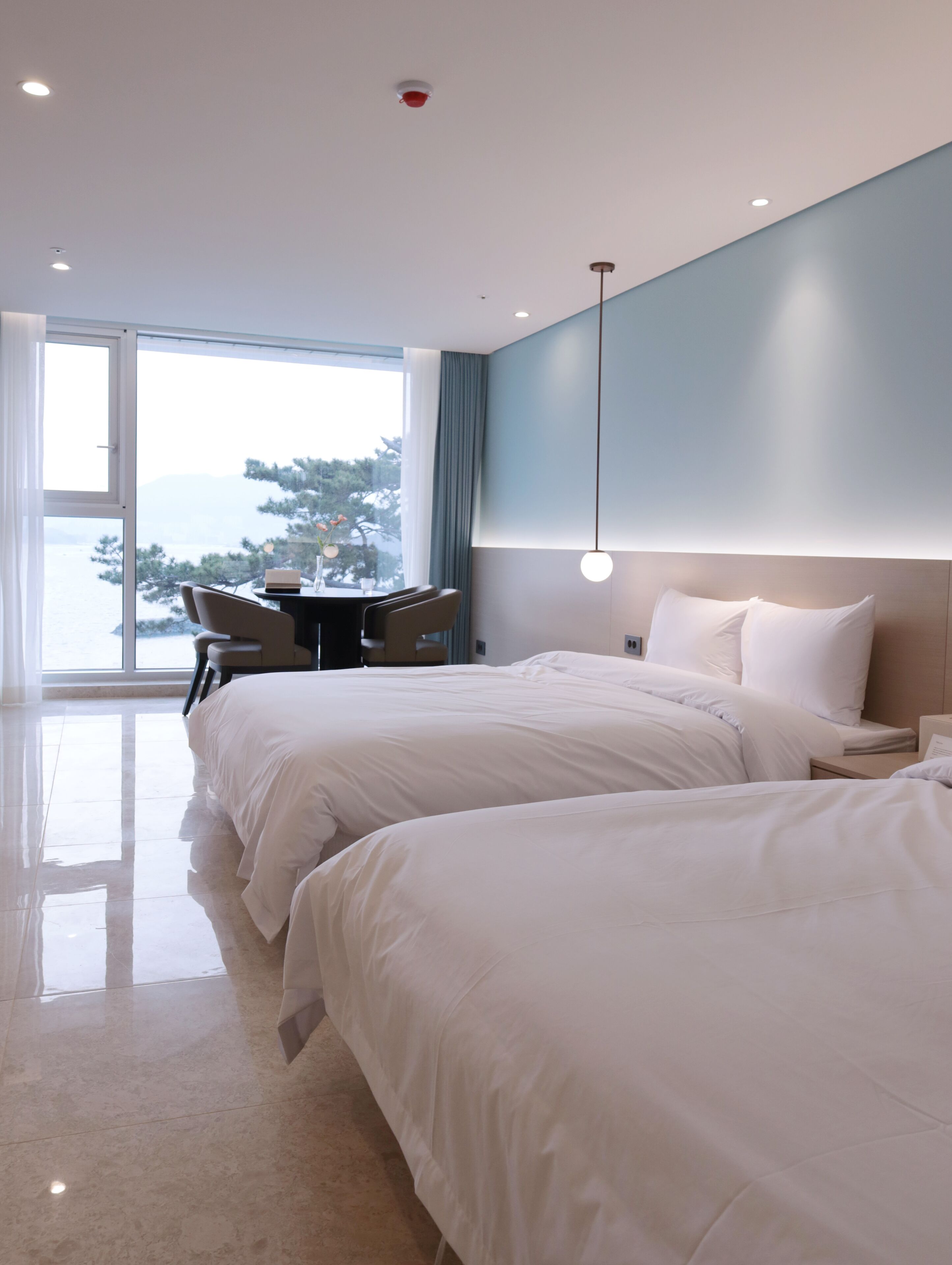 A modern hotel room with two beds, a dining area, and a view of the sea.