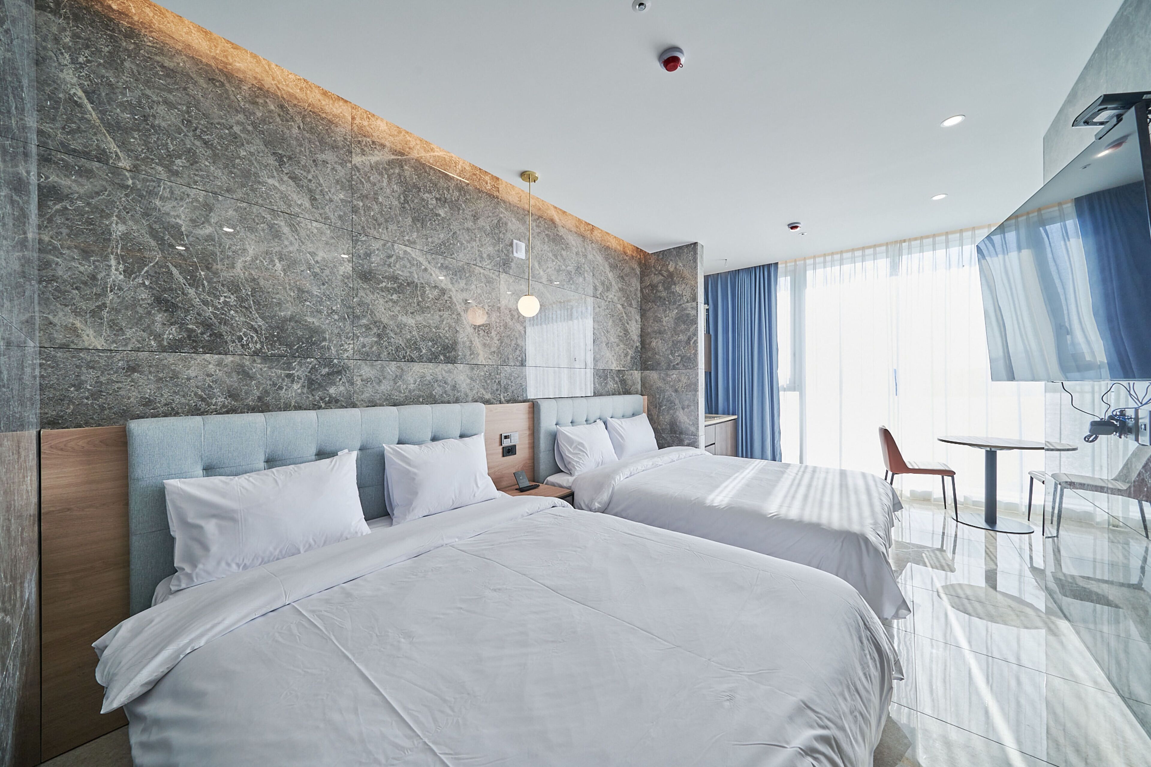 Ein modernes Hotelzimmer mit zwei Betten, Marmorwänden, einem Holzkopfende, einem kleinen Tisch mit Stühlen und einem großen Fenster mit Vorhängen.