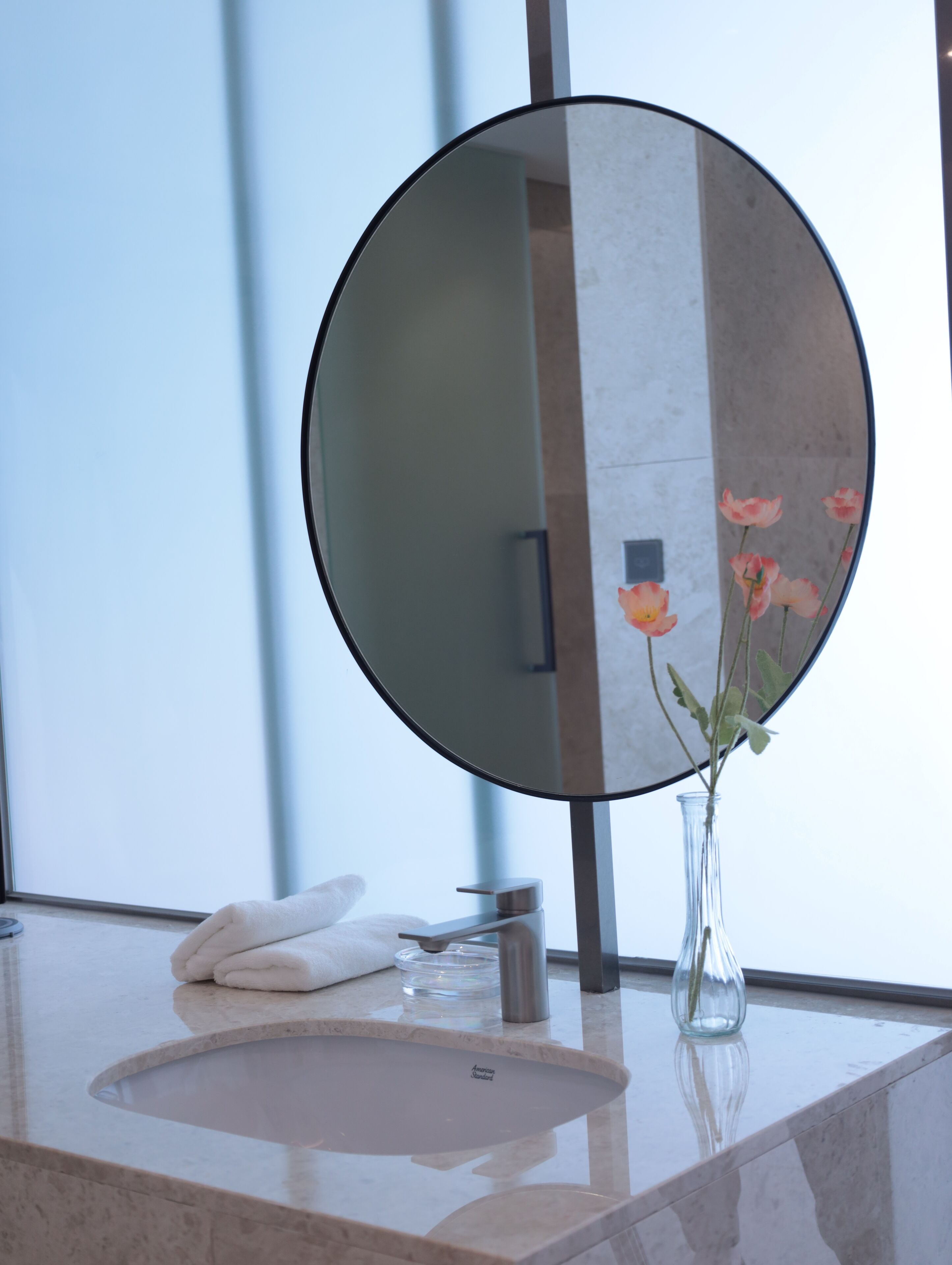 A bathroom with a round mirror, a sink, a vase with flowers, and a towel.