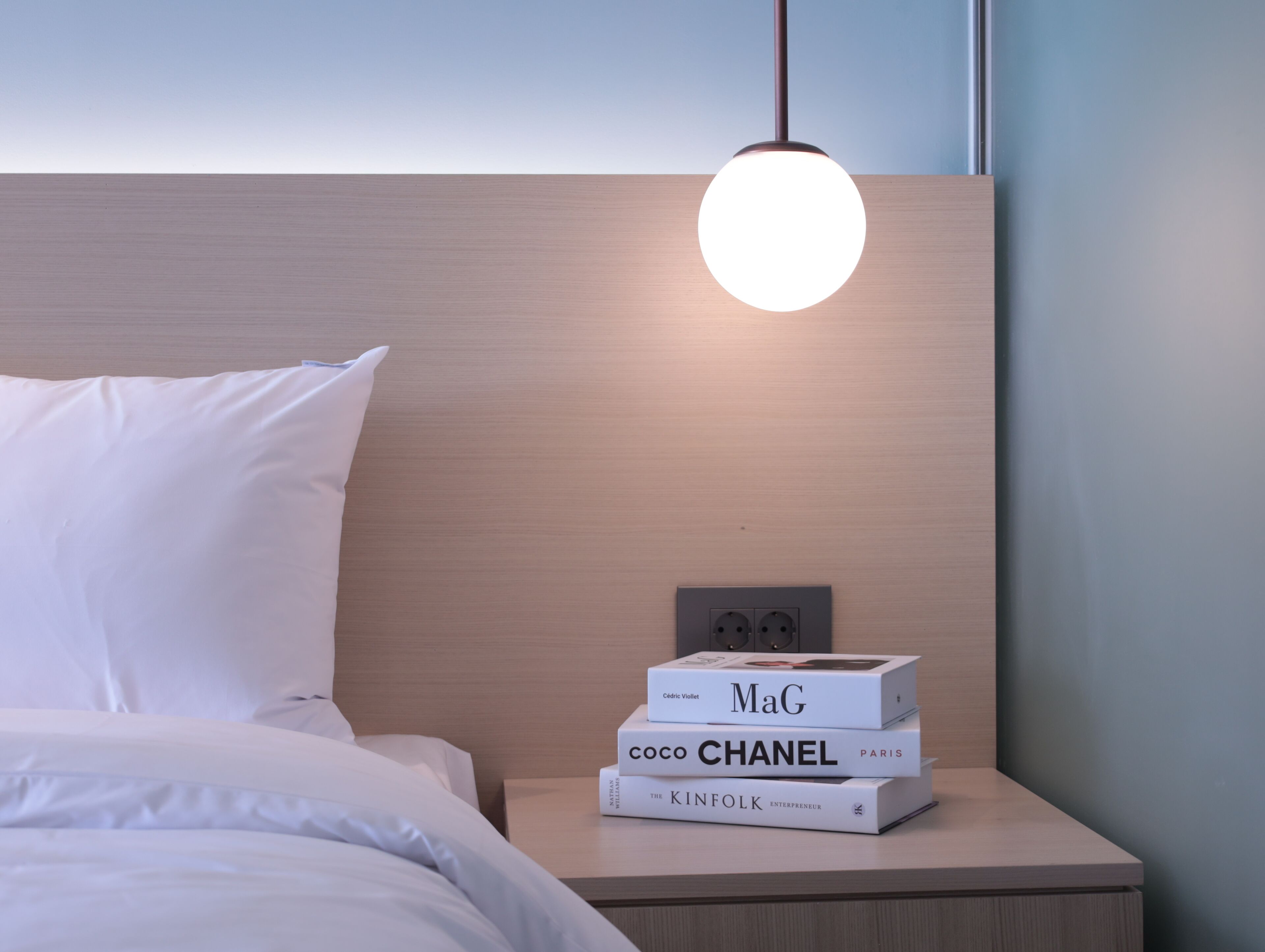 A neatly made bed with white linens, a wooden headboard, and a bedside table with a stack of books.