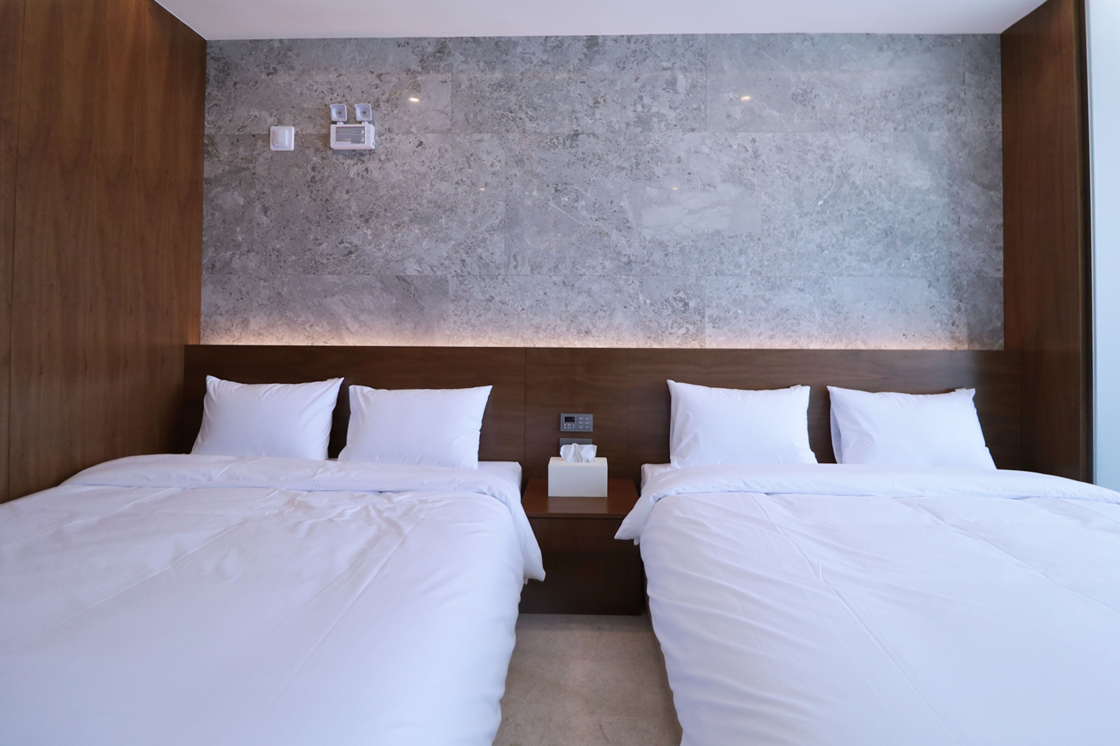A hotel room with two beds, a marble headboard, a nightstand with a tissue box, and a wall-mounted light fixture.