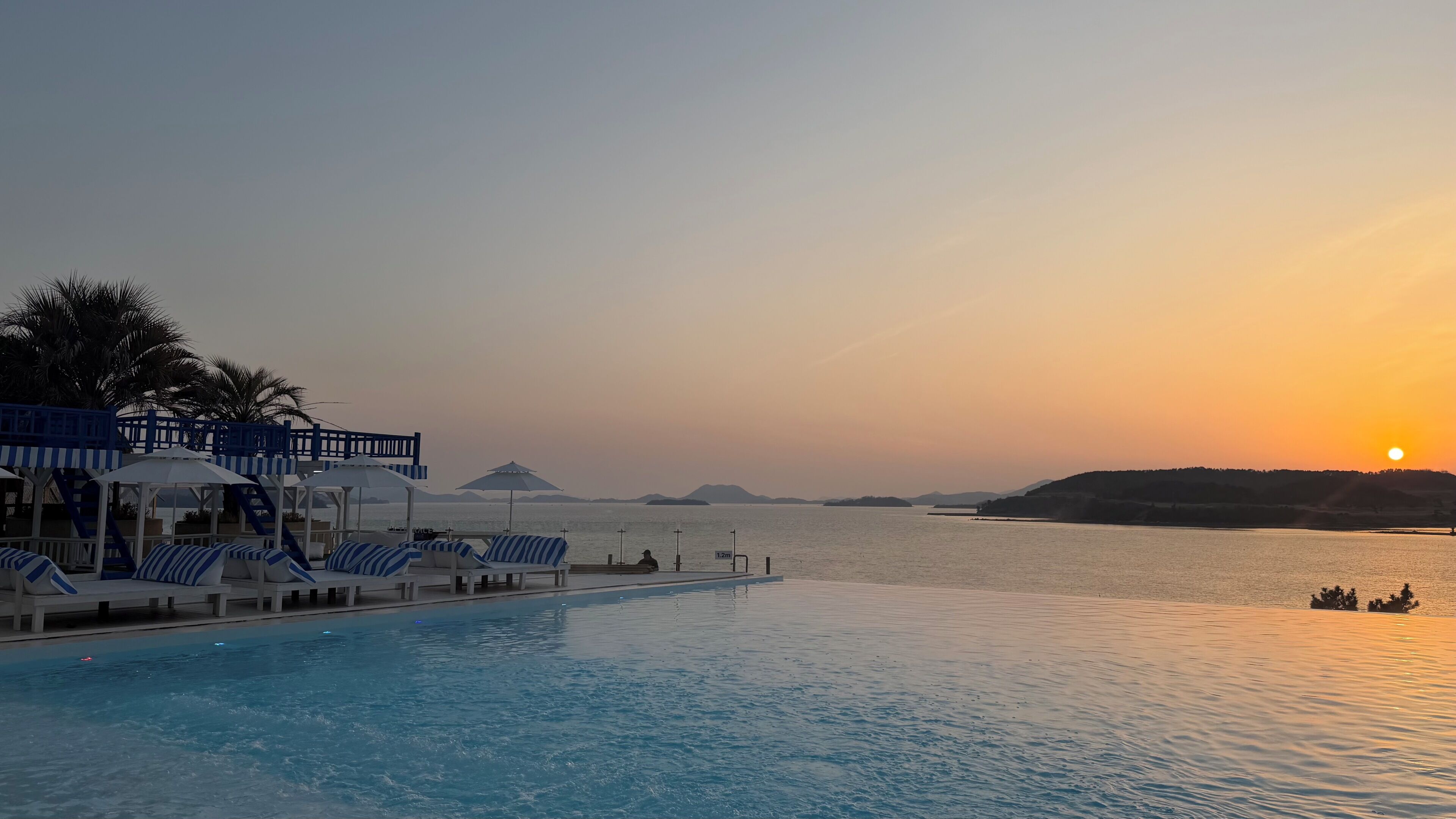 Zona junto a la piscina con sillones de descanso y sombrillas, con vista a una masa de agua tranquila al atardecer.