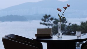A table with a book and a vase with flowers, overlooking a body of water and mountains. - Calacatta Hotel & Resort (Yeosu)