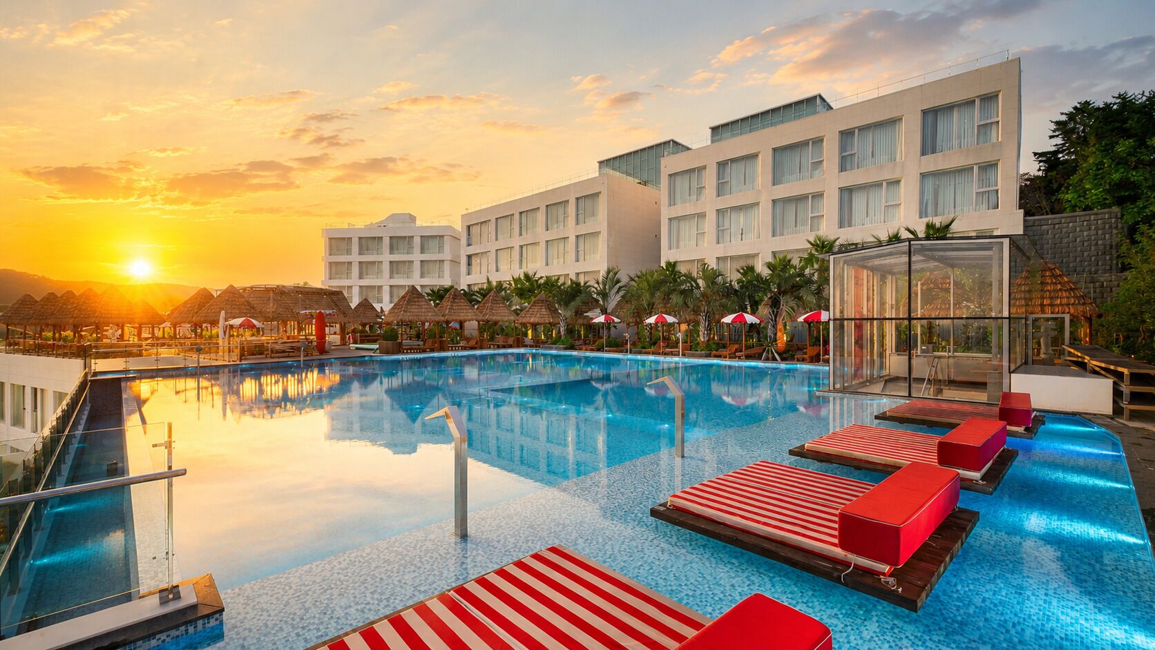 Ein Hotelpool mit freiem Blick auf den Sonnenuntergang, ein modernes mehrstöckiges Gebäude und eine Lounge mit rot gestreiften Liegen.