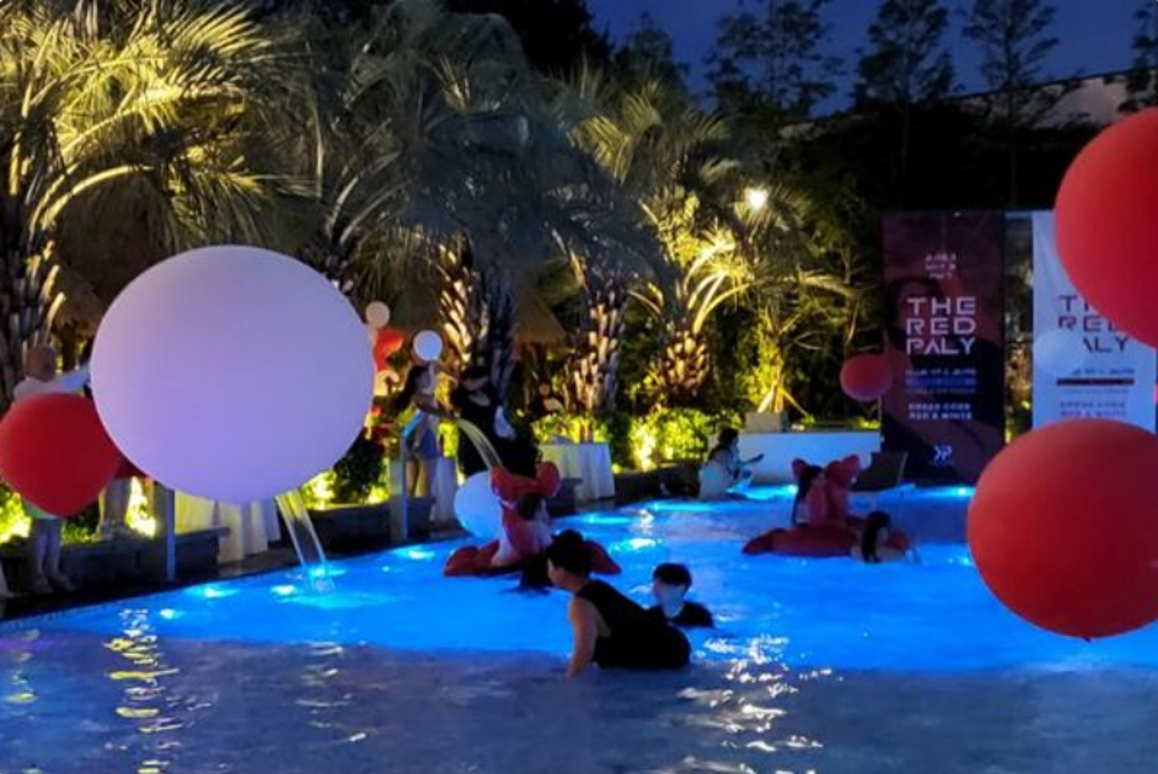 Zona de piscina con gente nadando y tomando el sol, rodeada de palmeras e iluminada con luces ambientales.