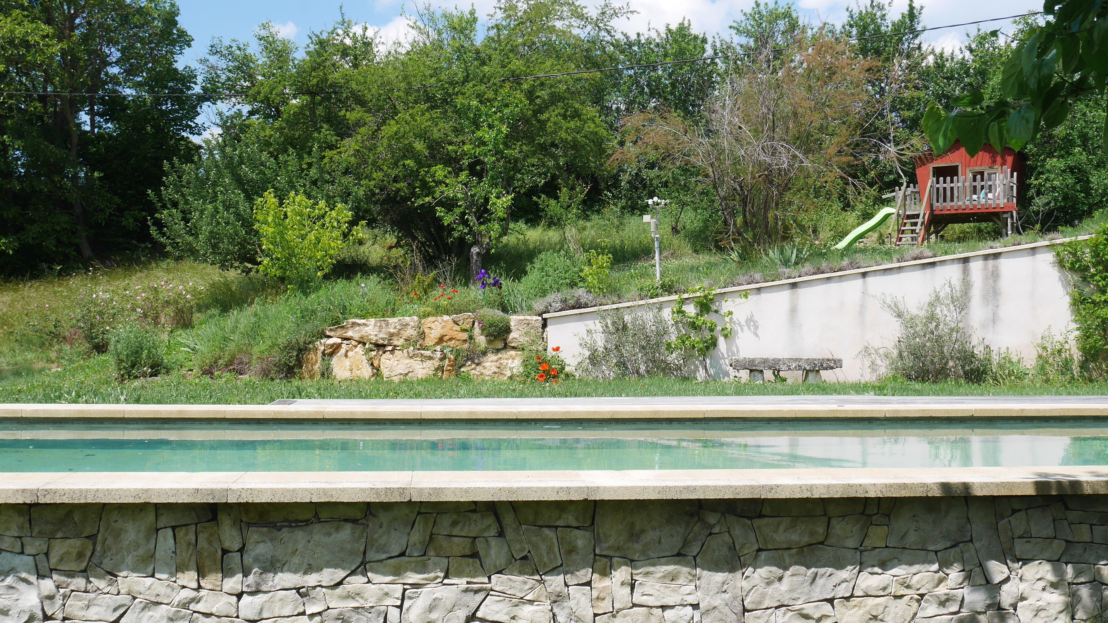 A swimming pool with a stone wall, a red playhouse, and surrounding greenery.