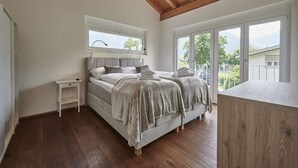 A bedroom with a large bed, a nightstand, a wooden ceiling, and a balcony with a view of greenery.