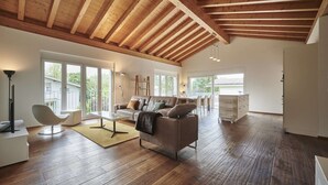 A spacious living room with wooden flooring, a high ceiling with exposed beams, a sofa, a coffee table, and a dining area with a table and chairs.