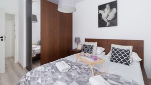 A modern bedroom with a wooden wardrobe, a bed with patterned bedding, a bedside table with a lamp, and a tray with items on it.