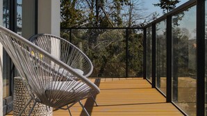 Ein Balkon mit zwei weißen Korbstühlen und einem Hocker mit geometrischem Muster, mit Blick auf eine bewaldete Landschaft.